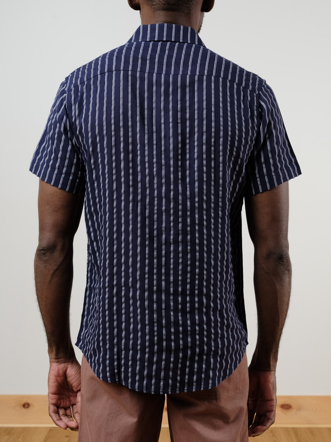 A person faces away from the camera, wearing the ID Menswear Drifter Linen Rayon Blend Shirt in navy blue and white stripes with light brown pants. The setting is indoors with a plain background and wooden floor.