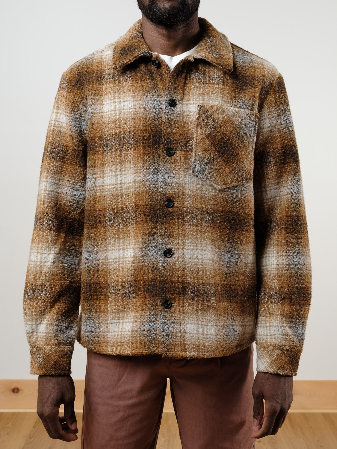 A person wears the !Solid Dronen Plaid Shirt Jacket by !SOLID, featuring brown, white, and gray plaid and a chest pocket, while standing indoors on a wooden floor against a plain light-colored background.