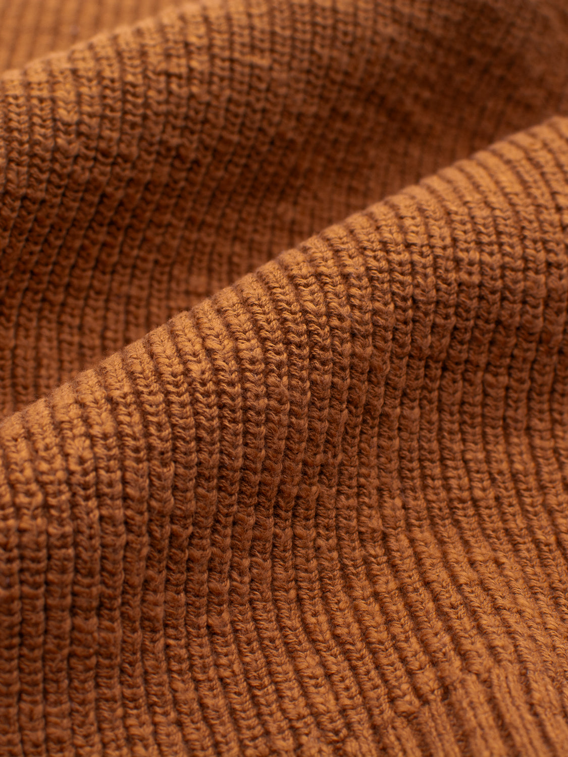 A close-up of the Dublin Slub Cotton Rolled Neck Sweater by ID Menswear shows its textured brown fabric with parallel ribbed patterns. The lighting highlights its woven structure, emphasizing its soft and inviting look, perfect for cooler weather.