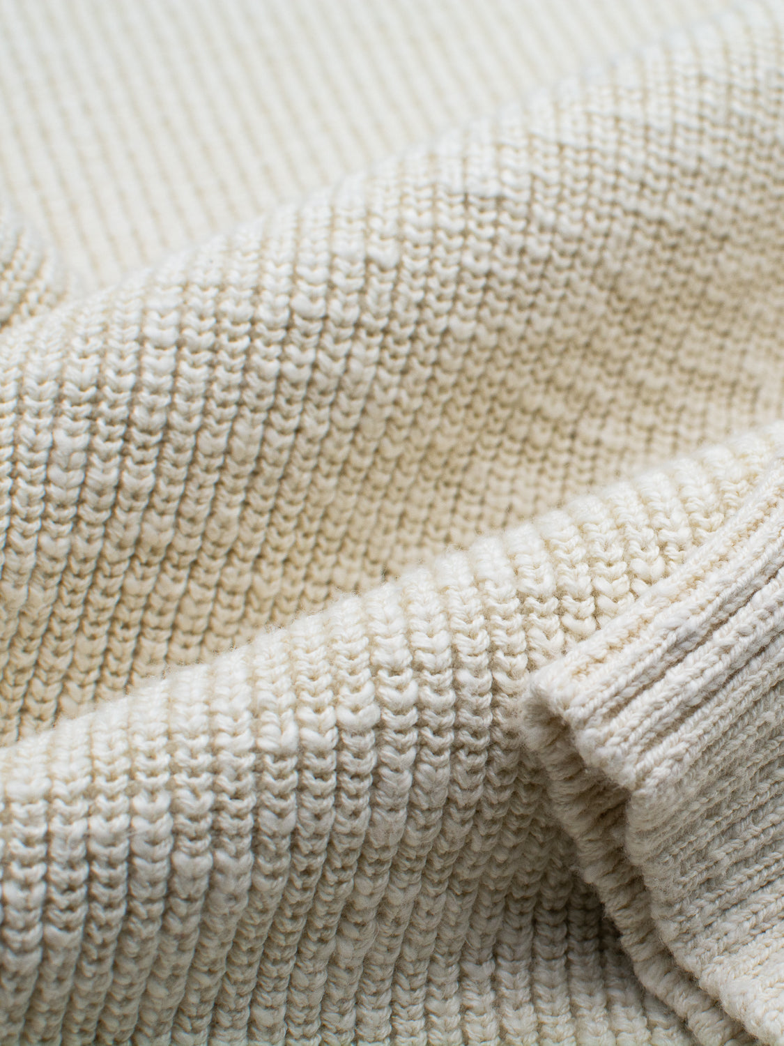 Close-up of the Dublin Slub Cotton Rolled Neck Sweater by ID Menswear in a ribbed, cream-colored knit. The fabrics texture and weave offer a soft, cozy feel, perfect for cooler weather, with gentle folds showcasing its thickness and warmth.
