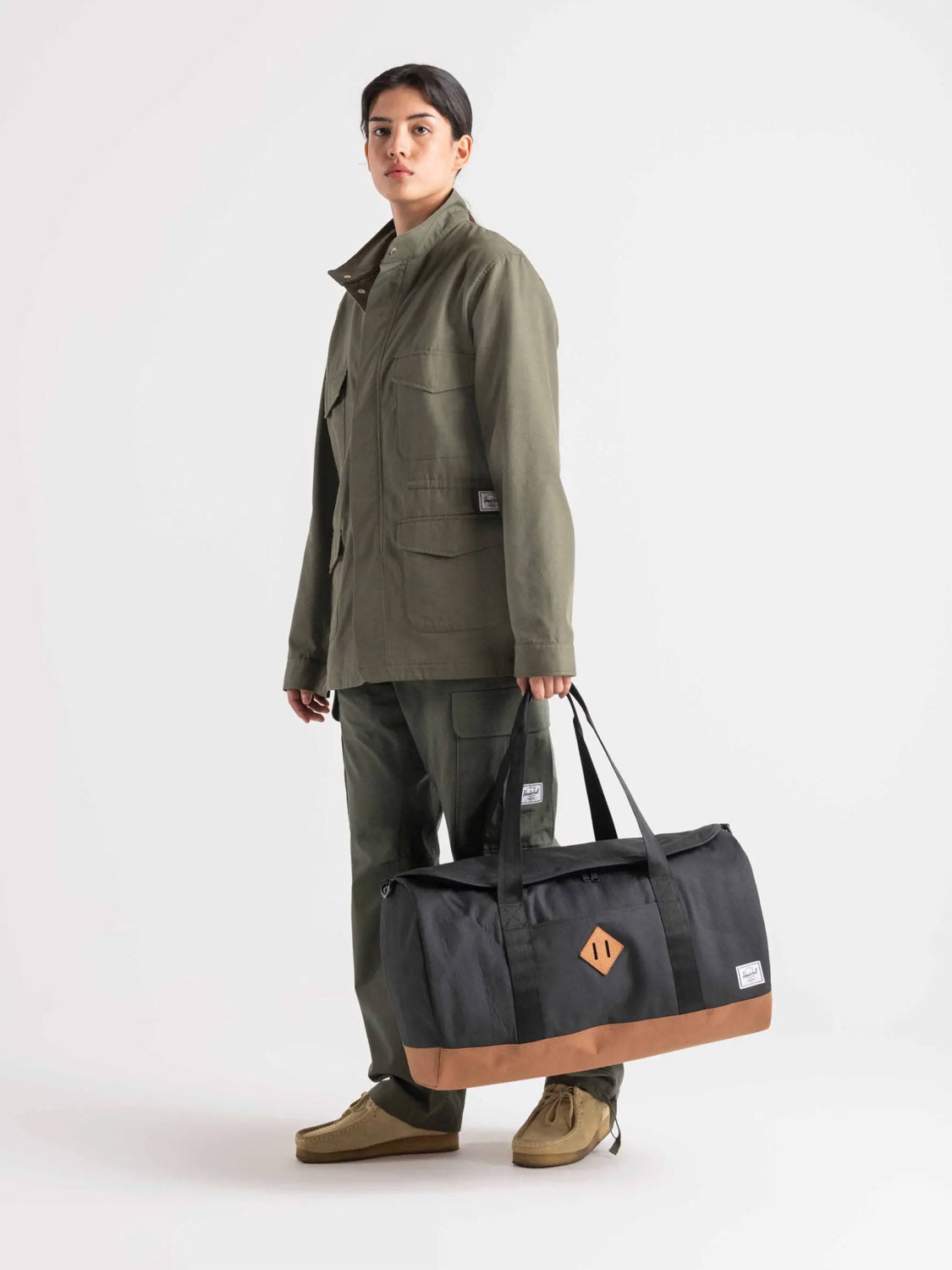 A person in an olive green utility jacket and pants stands facing forward, holding the Herschel Heritage™ Duffle - 40L, made from recycled polyester, against a plain white background.