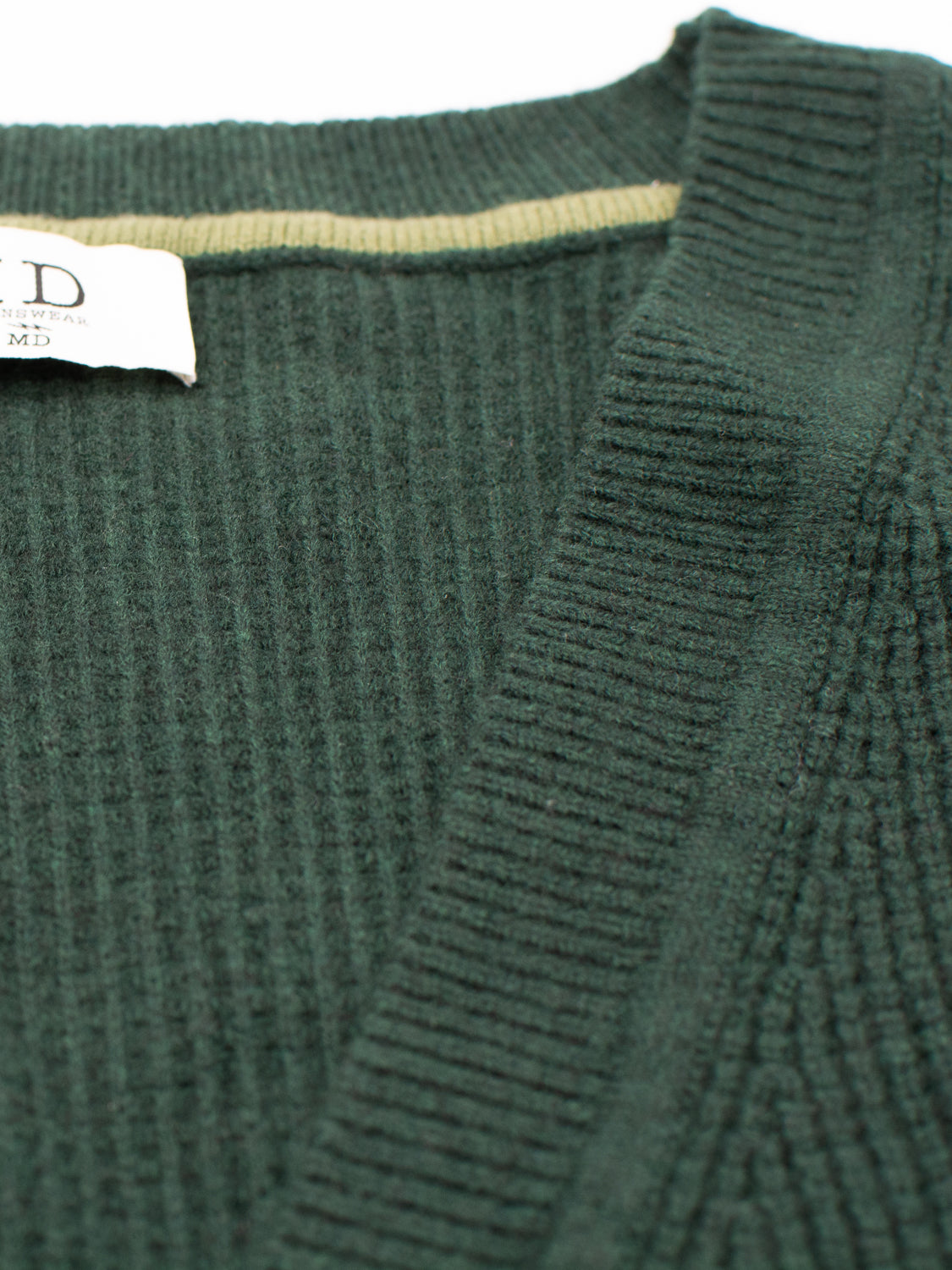 Close-up of the Dundee Waffle Knit Merino Cardigan by ID Menswear, featuring dark green ribbed knit fabric, a V-neckline, and a white tag visible at the collar that showcases the detailed stitching and texture.