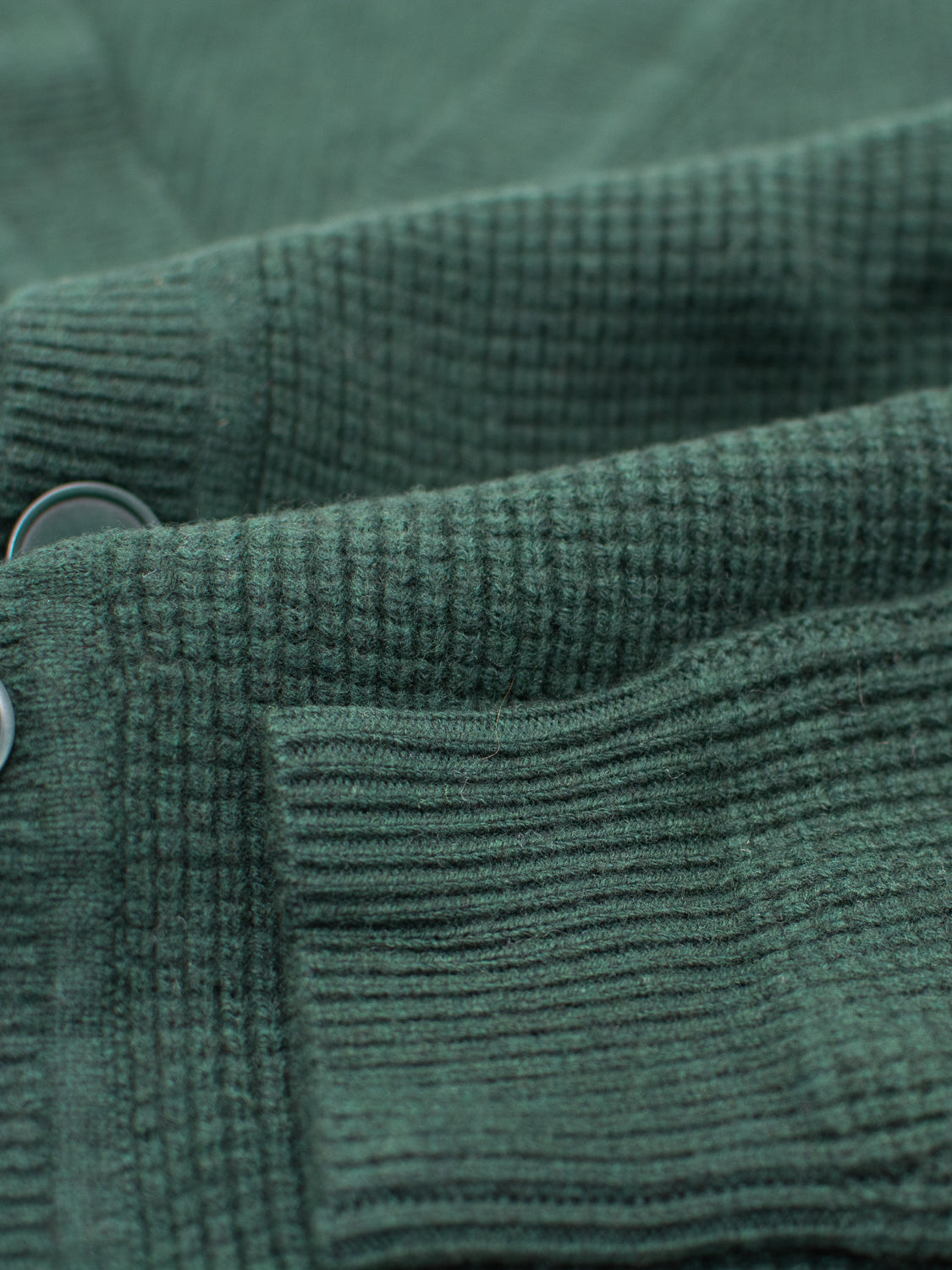 Close-up of textured green Dundee Waffle Knit Merino Cardigan by ID Menswear, highlighting ribbed knit patterns and two round buttons partially visible on the left.