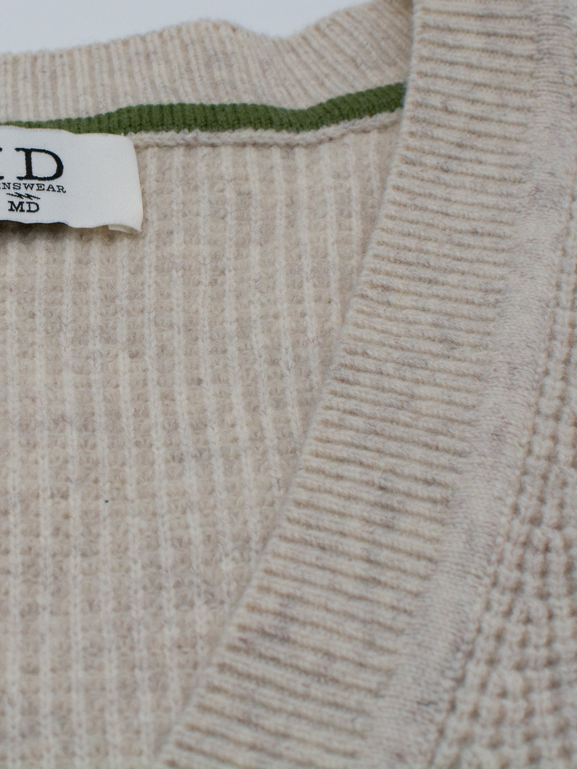 Close-up of the Dundee Waffle Knit Merino Cardigan by ID Menswear in beige, featuring a thick collar, ribbed knit texture, green inner trim, and a white tag with "MD" and "ID Menswear" at the neckline.