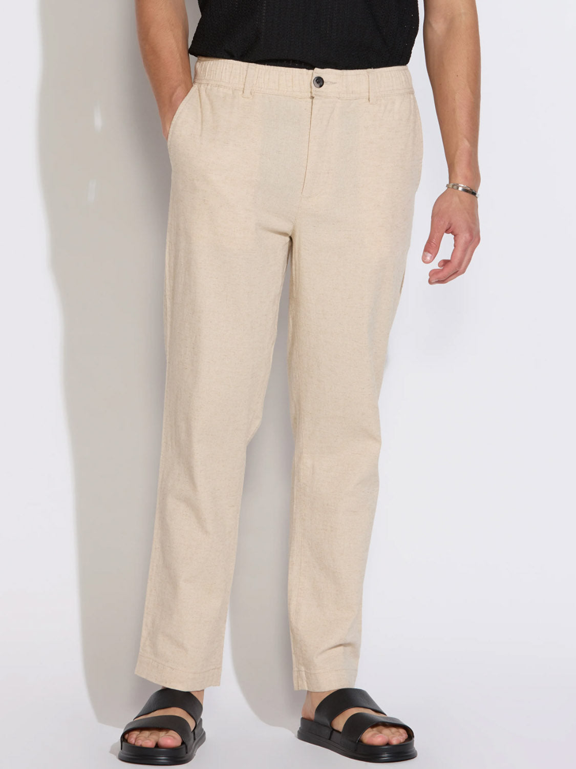 A person wearing Civil Society’s Dune Classic Straight Fit Trouser in light beige stands with hands in pockets, paired with black sandals and a black top. Only the lower body is shown against a plain white background.