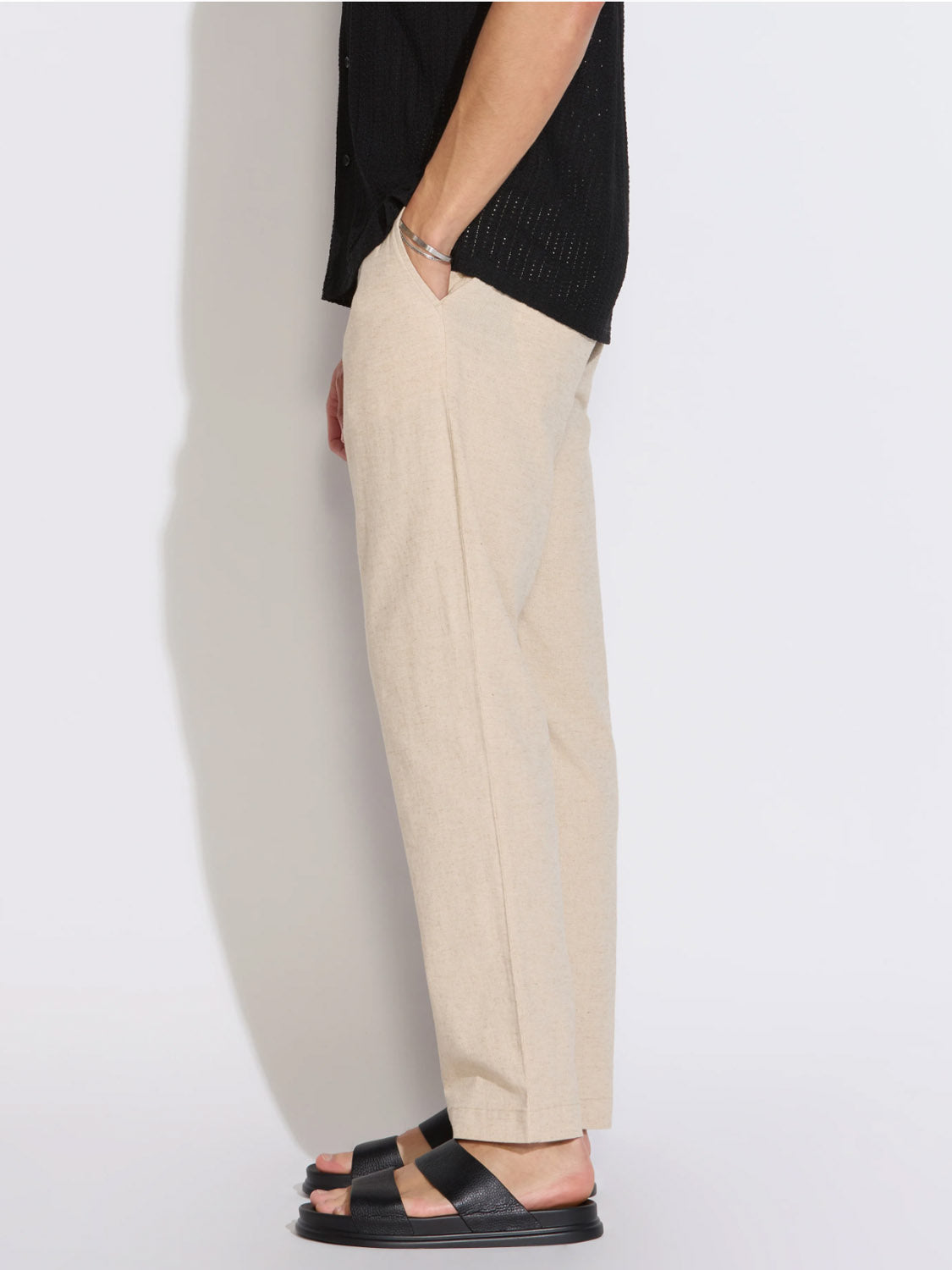 A person stands in profile against a white background, showing only their lower half, dressed in Civil Society's Dune Classic Straight Fit Trouser in beige, paired with black sandals and a bracelet.