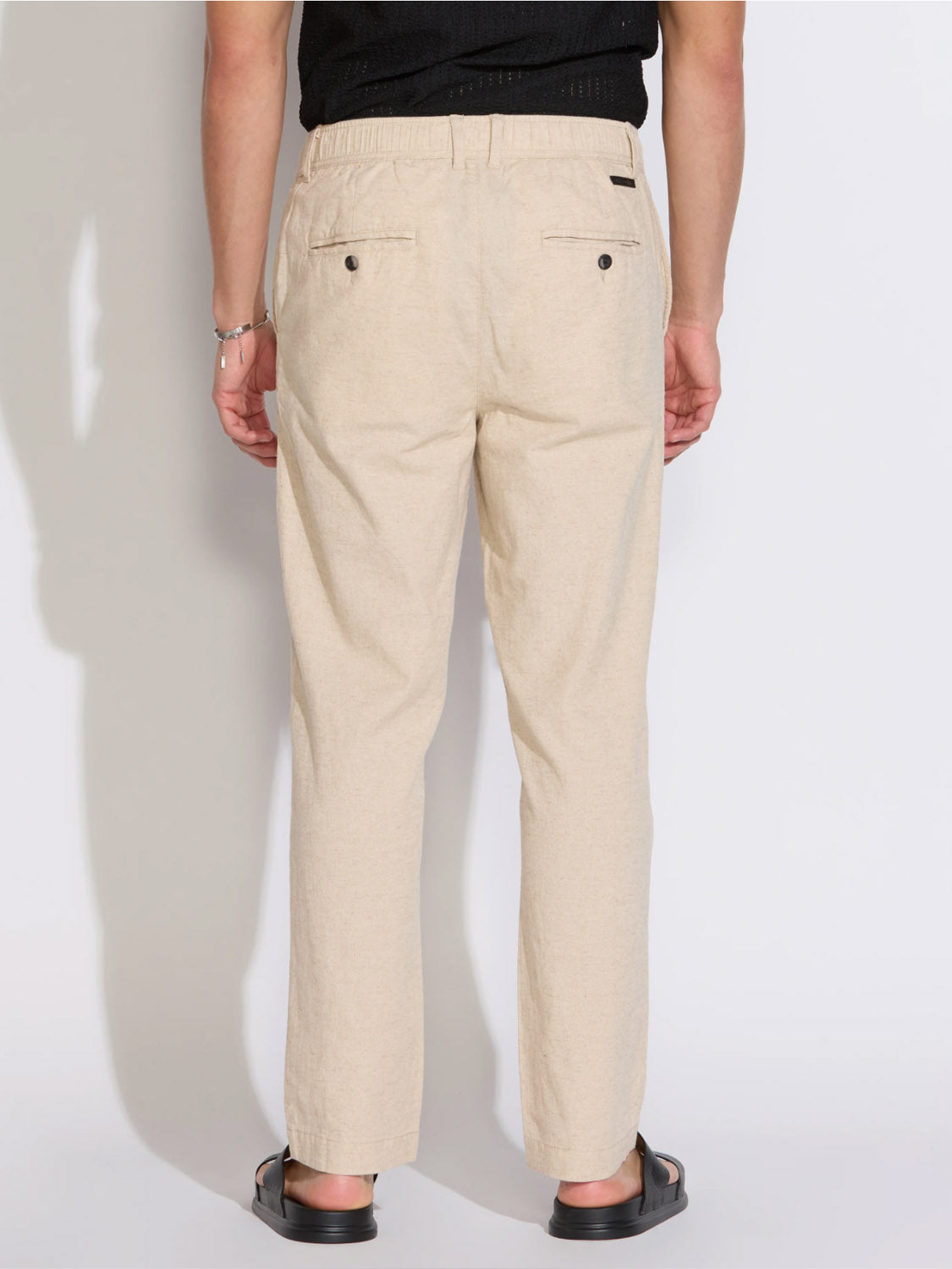 A person stands facing away wearing Civil Society’s Dune Classic Straight Fit Trouser in light beige, paired with a black shirt, black sandals, and a bracelet; the trousers feature two back pockets with buttons.