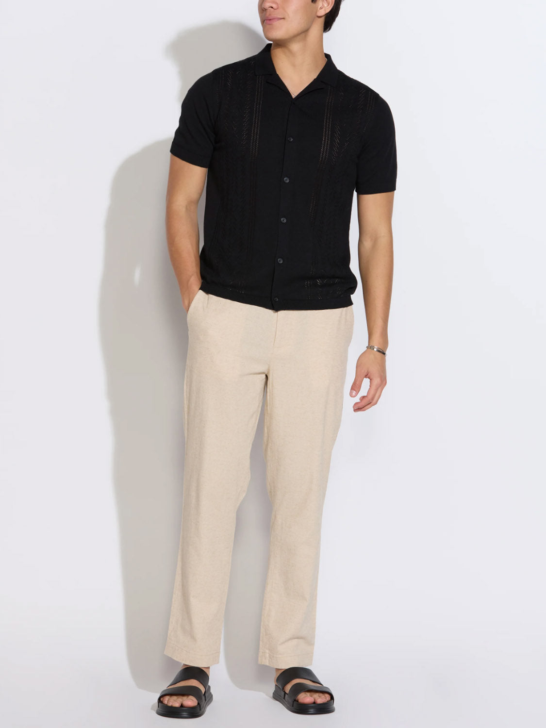 Wearing a short-sleeve black button-up shirt, Civil Society Dune Classic Straight Fit Trouser in light beige, and black sandals, a person stands with hands in pockets against a plain white background.