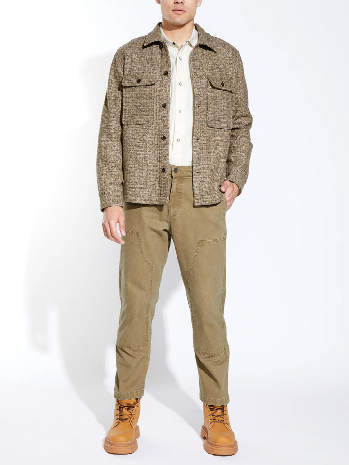 A man poses against a white background wearing Civil Society's Durbin Relaxed Fit Shirt Jacket, a beige shirt, olive relaxed fit pants, and tan lace-up boots. His face is partially out of view.