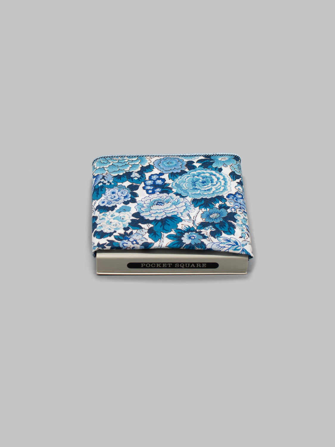 The Liberty™ Elysian Pocket Square by ID Menswear, crafted from blue and white floral Liberty of London Tana Lawn cotton, is neatly folded in a beige box labeled POCKET SQUARE on a plain grey background.