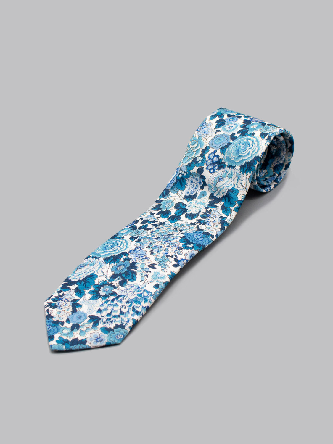 The ID Menswear Liberty of London Tana Lawn™ Elysian Tie, featuring a blue and white floral pattern and handmade in Brooklyn from fine cotton, is shown rolled at one end on a plain grey background to showcase its elegant design.