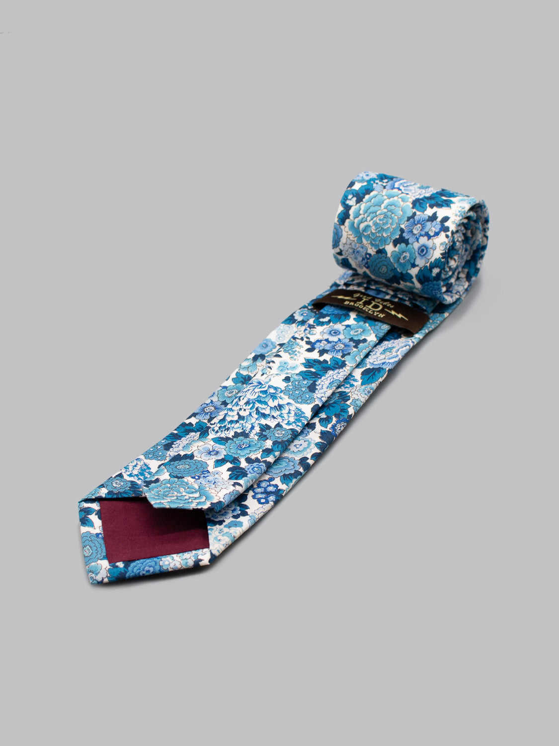 The Liberty of London Tana Lawn™ Elysian Tie by ID Menswear features a blue and white floral pattern on gray, handcrafted in Brooklyn from fine cotton, with a maroon-tipped underside and black label near the roll.
