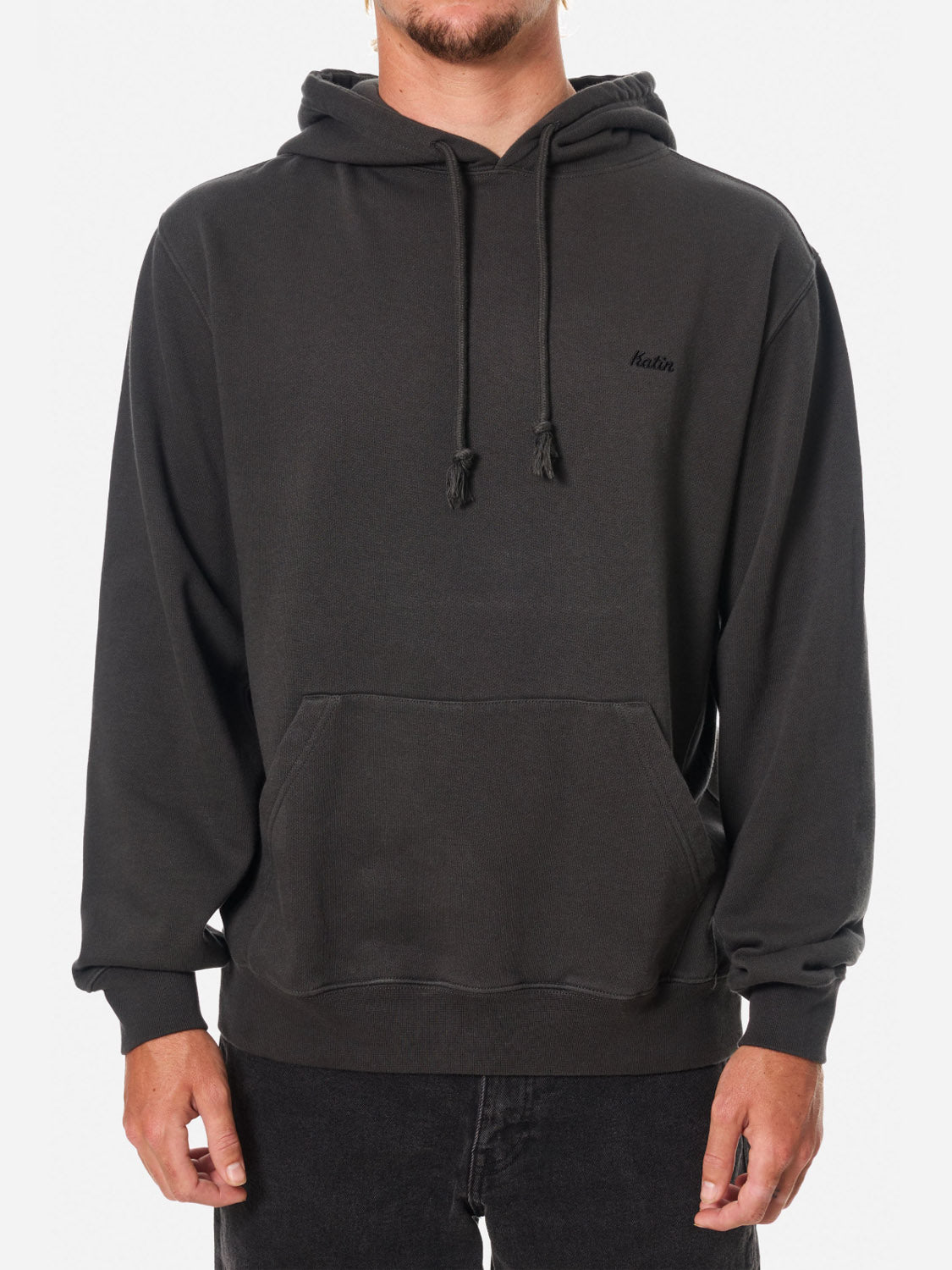 A person wears the Katin Embroidered Hoodie, featuring heavyweight fleece, subtle left-chest text, front pocket, and drawstrings, paired with dark pants and posing against a white background.