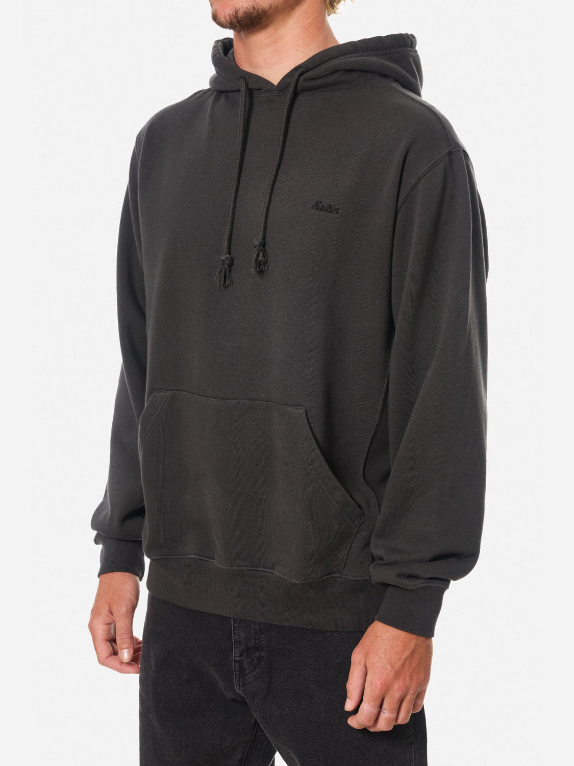 A person stands against a white background wearing the Katin Embroidered Hoodie, a loose-fitting, garment-washed black hoodie with embroidery, front pocket, and black drawstrings, paired with black jeans.