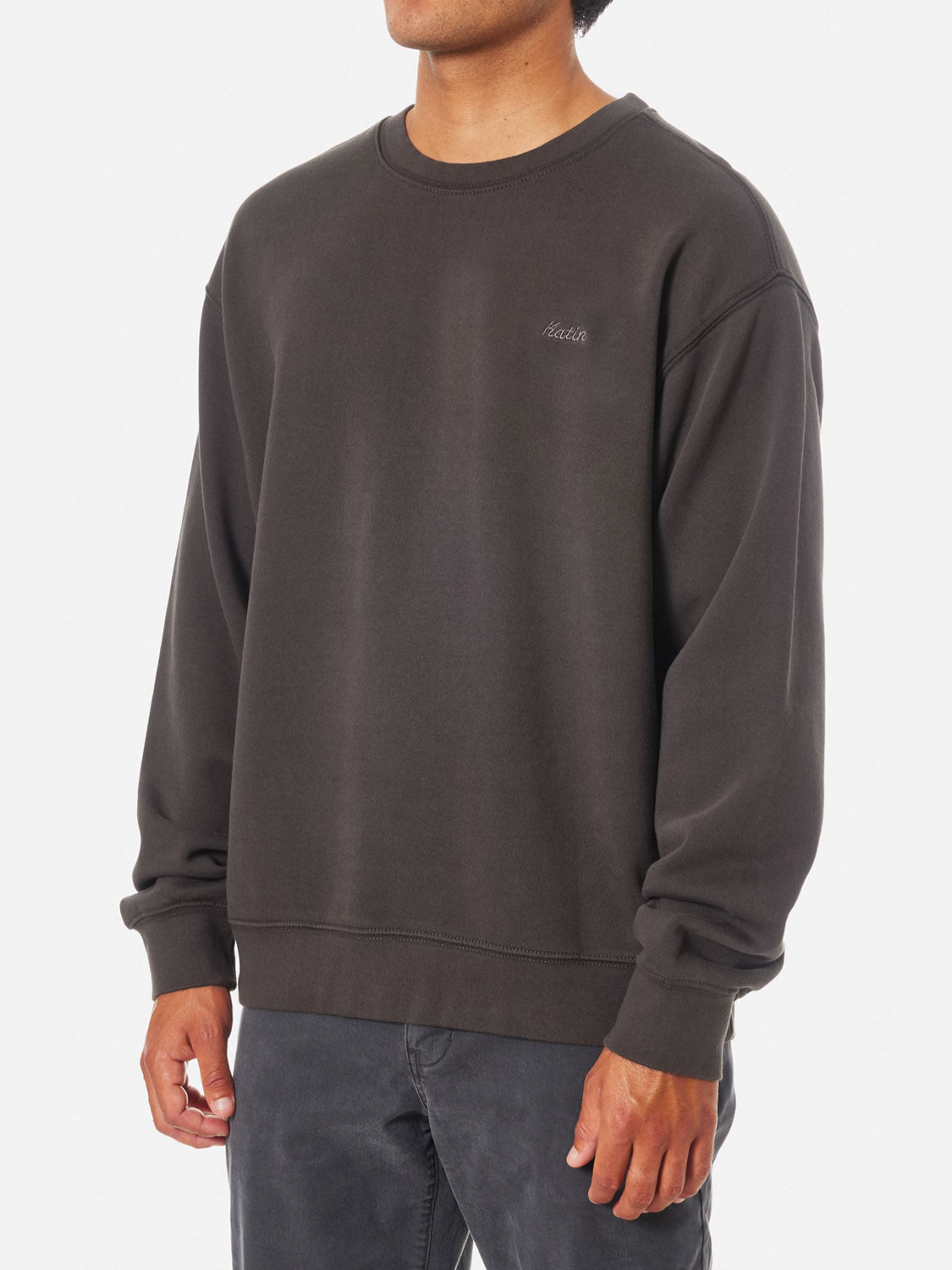 A person in a loose-fitting Katin Embroidered Crewneck, featuring dark brown fabric, long sleeves, and subtle logo embroidery on the left chest, paired with dark pants, stands against a plain white background.