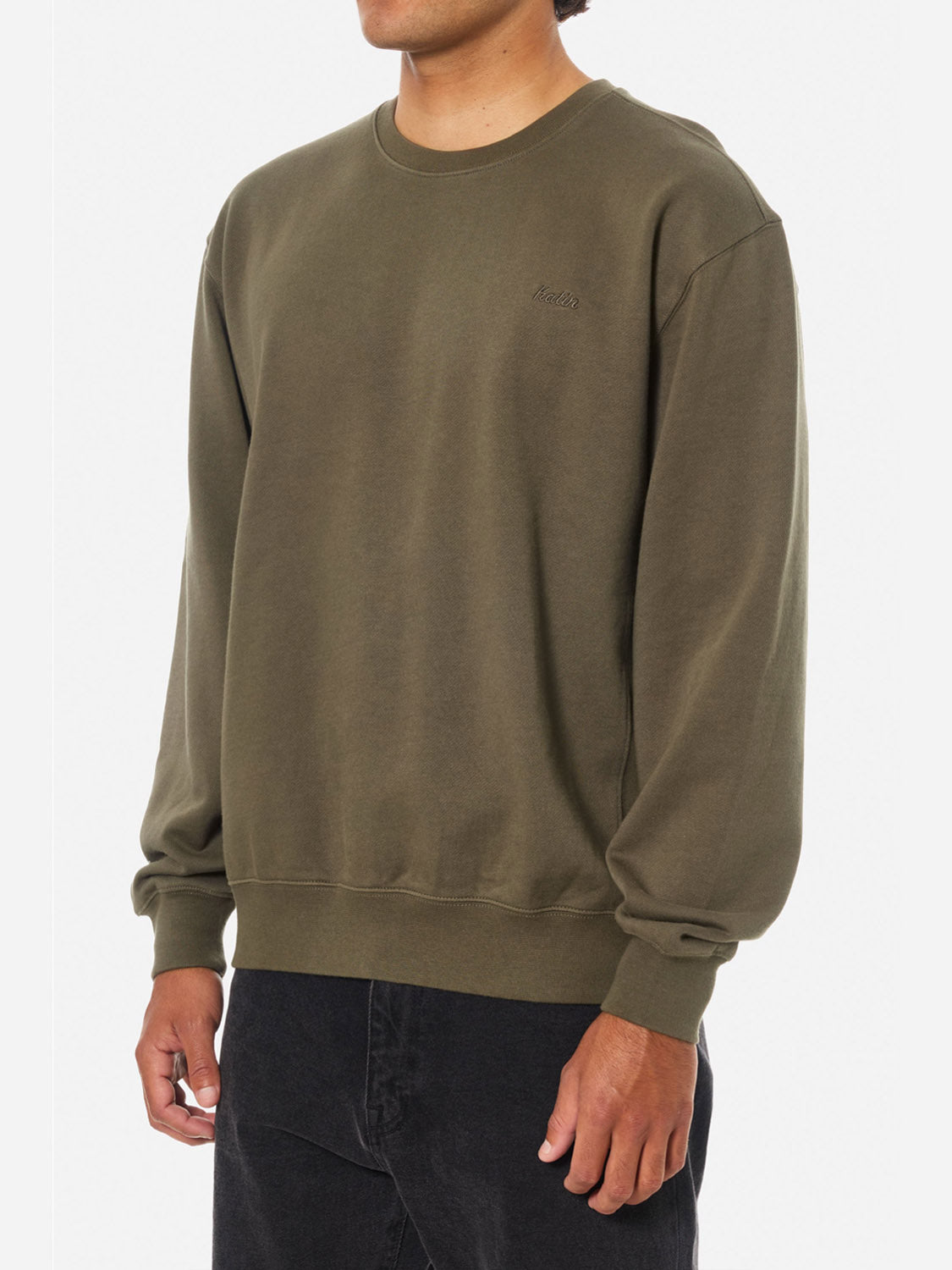 A person wears the Katin Embroidered Crewneck in olive green with long sleeves and black pants, standing against a plain white background.