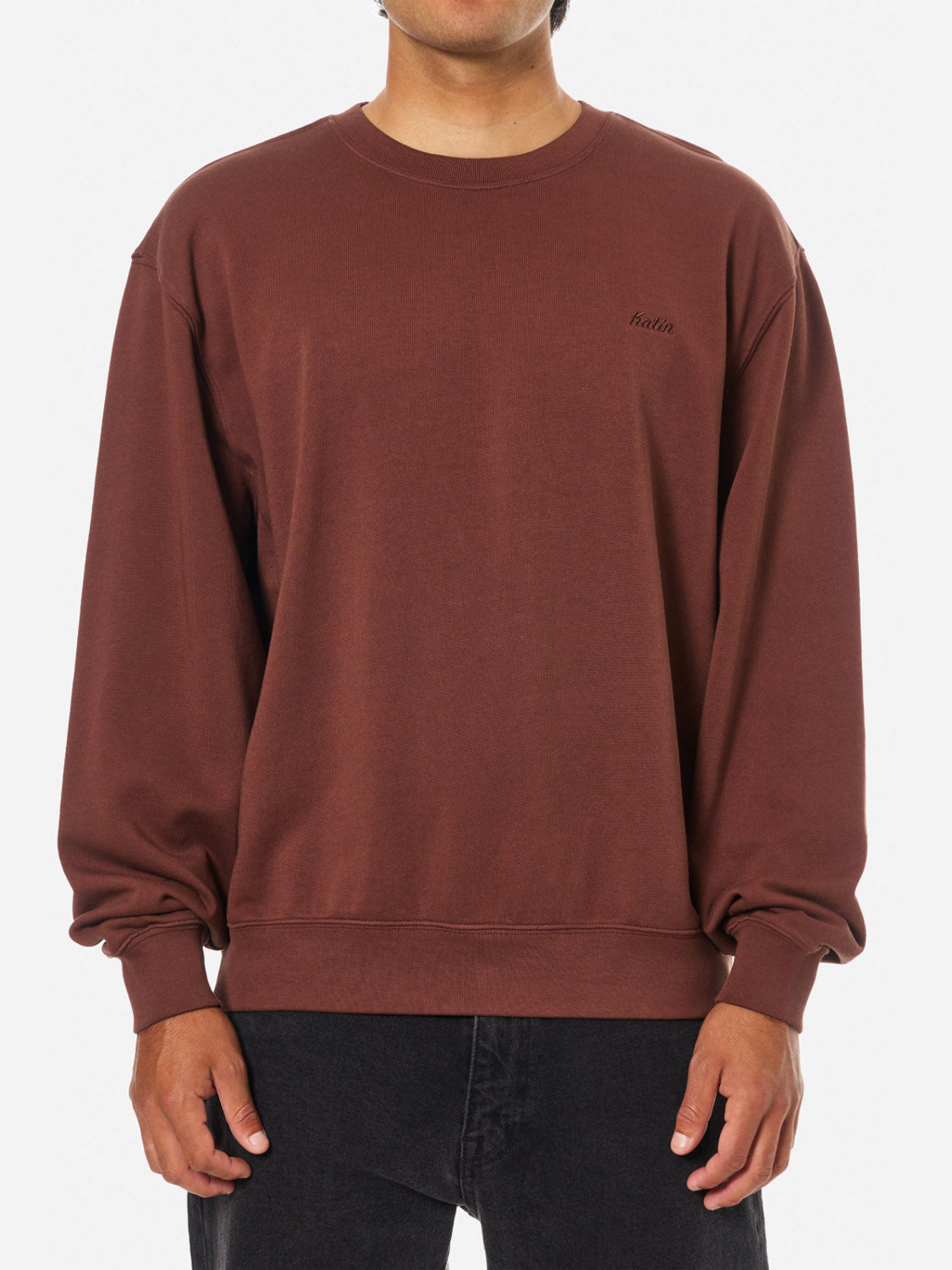 A person stands against a plain white background wearing the Katin Embroidered Crewneck in brown, featuring subtle text on the left chest, paired with black pants.