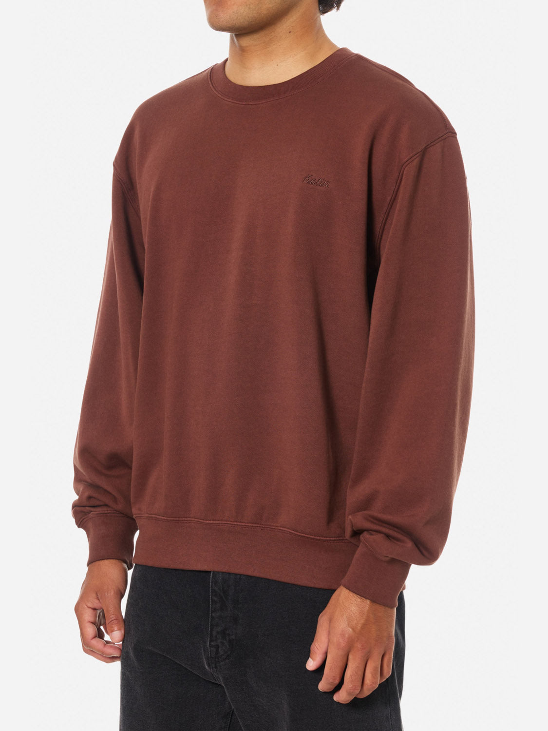 A person wearing Katin's Embroidered Crewneck, a loose-fitting, long-sleeve brown sweatshirt, paired with black pants and standing against a plain white background.