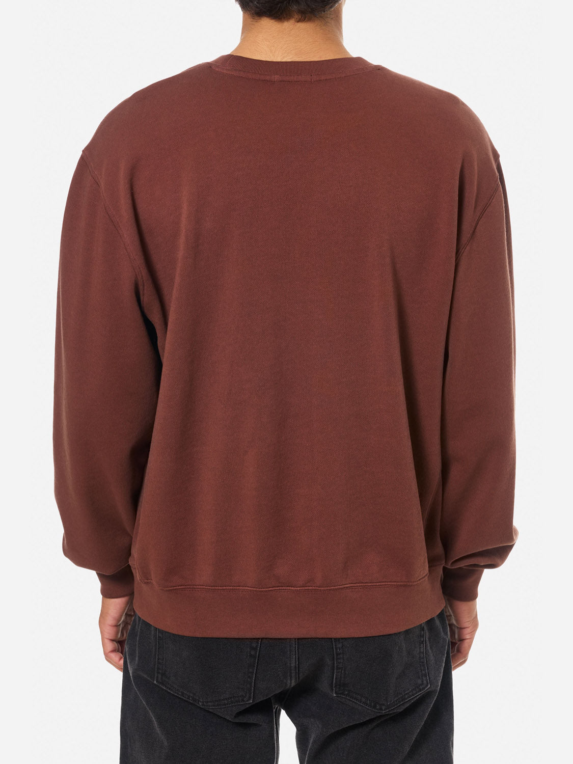 A person is shown from behind wearing Katin’s Embroidered Crewneck—a loose-fitting, long-sleeve, vintage wash brown sweatshirt made of soft fleece—paired with black jeans, all set against a plain white background.