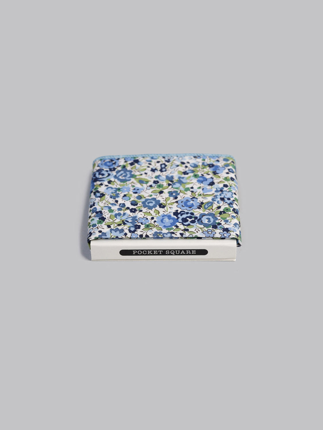 A folded Liberty™ Emma Pocket Square by ID Menswear, featuring blue and white florals, rests in a white box labeled POCKET SQUARE against a light grey background.