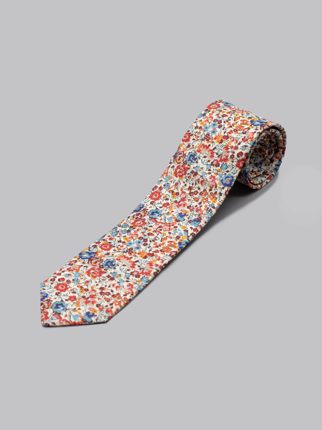 A rolled-up Liberty of London Tana Lawn™ Emma Rouge tie by ID Menswear, handmade in Brooklyn with a colorful red, blue, and orange floral pattern on white cotton, shown on a light gray surface.