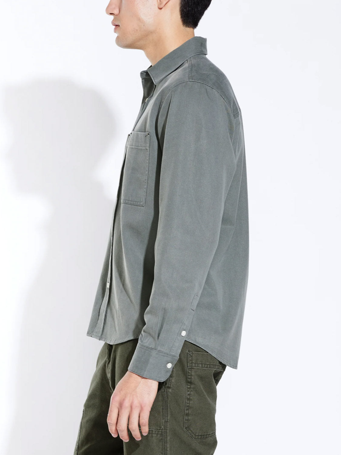 A person in profile wears the Civil Society Ernst Relaxed Fit Long Sleeve Shirt in olive green with a chest pocket and matching pants, evoking classic American workwear style against a plain white background.