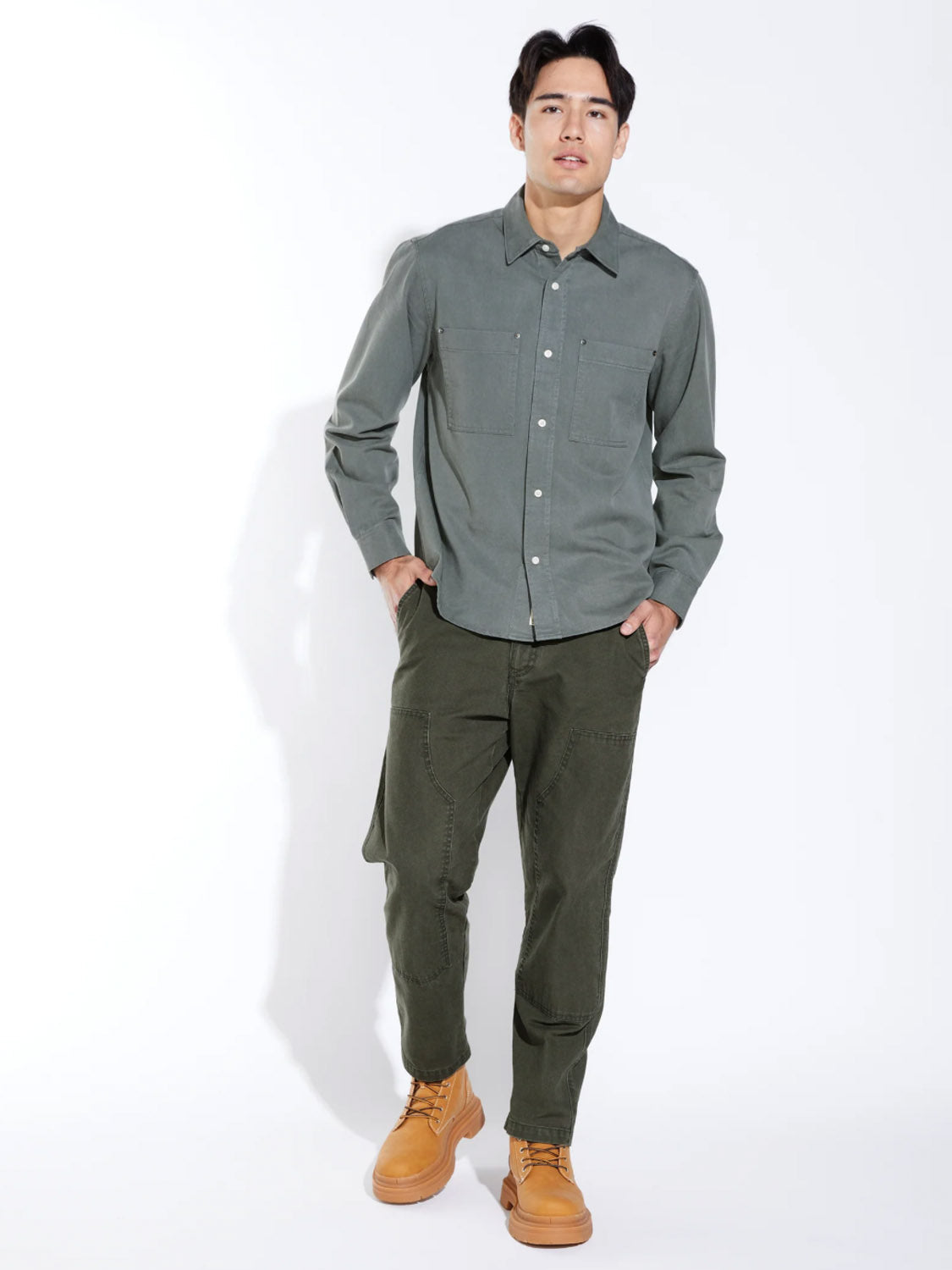 A man stands against a white background wearing the Civil Society Ernst Relaxed Fit Long Sleeve Shirt and matching pants with tan lace-up boots. His hands are in his pockets and he looks forward, embodying classic American workwear style.