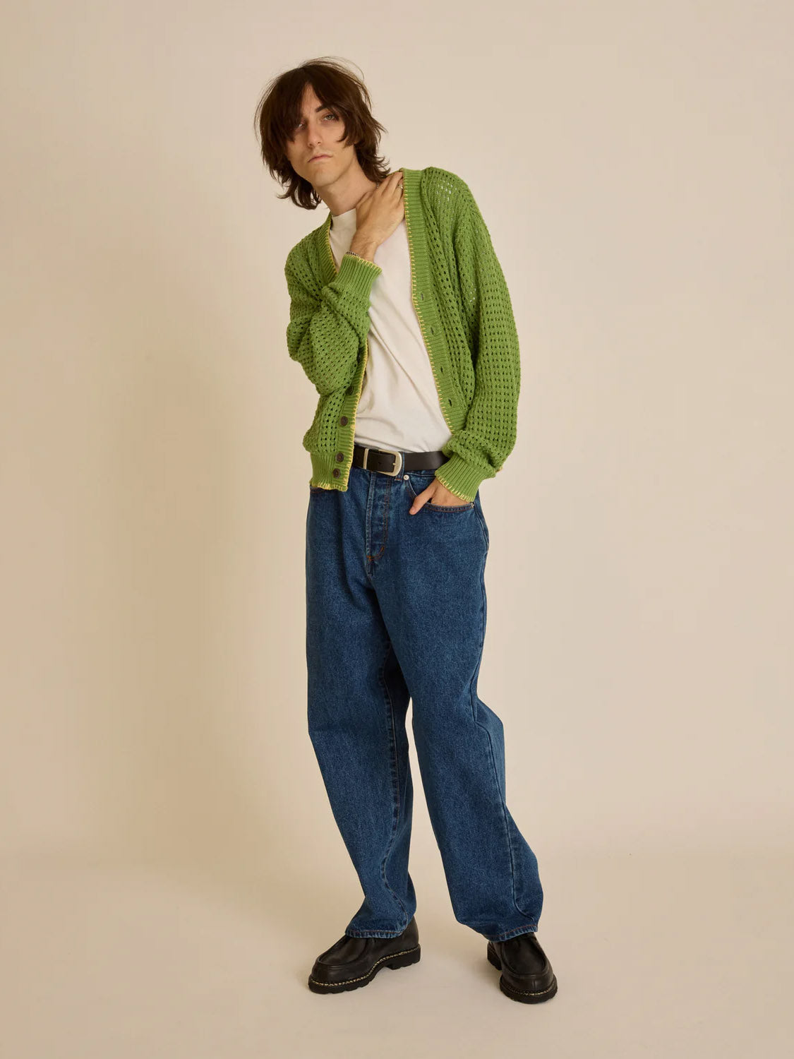 A person with medium-length hair is wearing an Obey Ethan Cardigan over a white t-shirt, blue jeans, and black shoes. They stand against a plain beige background, one hand on their chest and the other in a pocket.