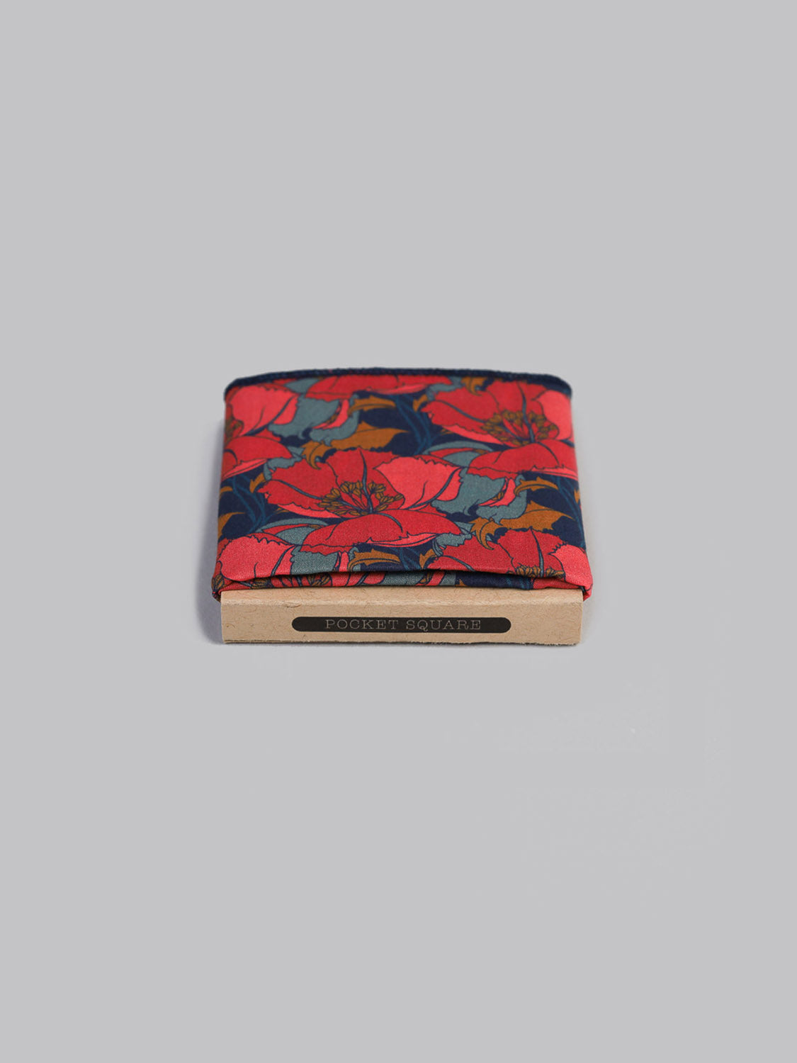 A folded Liberty™ Eustacia Pocket Square by ID Menswear, handmade in Brooklyn from soft Tana Lawn cotton, shows a bold red floral pattern with green leaves, displayed atop a beige box on a light gray background.