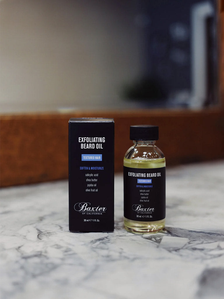 A bottle of Baxter Exfoliating Beard Oil sits beside its black box on a marble countertop. Designed for textured hair, it softens and moisturizes with key ingredients salicylic acid and jojoba oil.