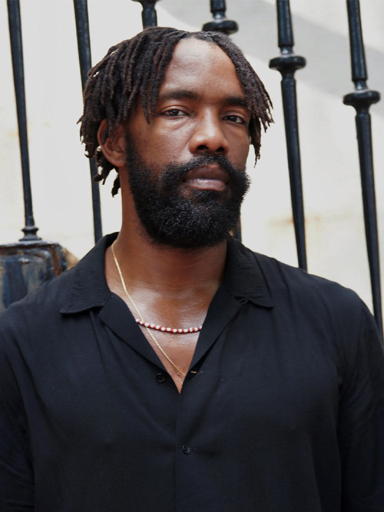 A man with a textured beard and short dreadlocks wears a black shirt and layered necklaces, standing outdoors. He uses Baxter’s Exfoliating Beard Oil for a well-groomed look.