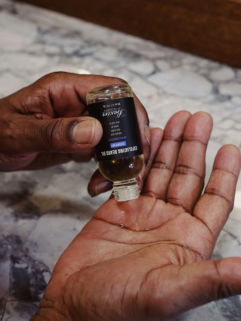 A person pours Baxter Exfoliating Beard Oil with salicylic acid from a small bottle into their palm over a marble countertop, perfect for caring for textured hair.