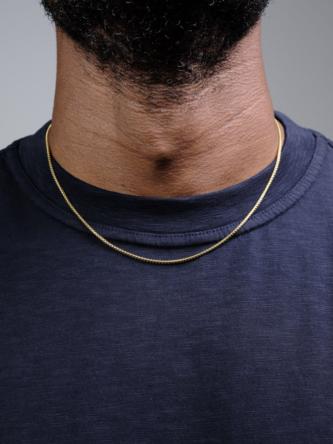 A close-up of a person wearing the ID Menswear Ezra 35 Box Chain Necklace over a dark blue crew-neck t-shirt, with only the lower half of their face and neck visible.