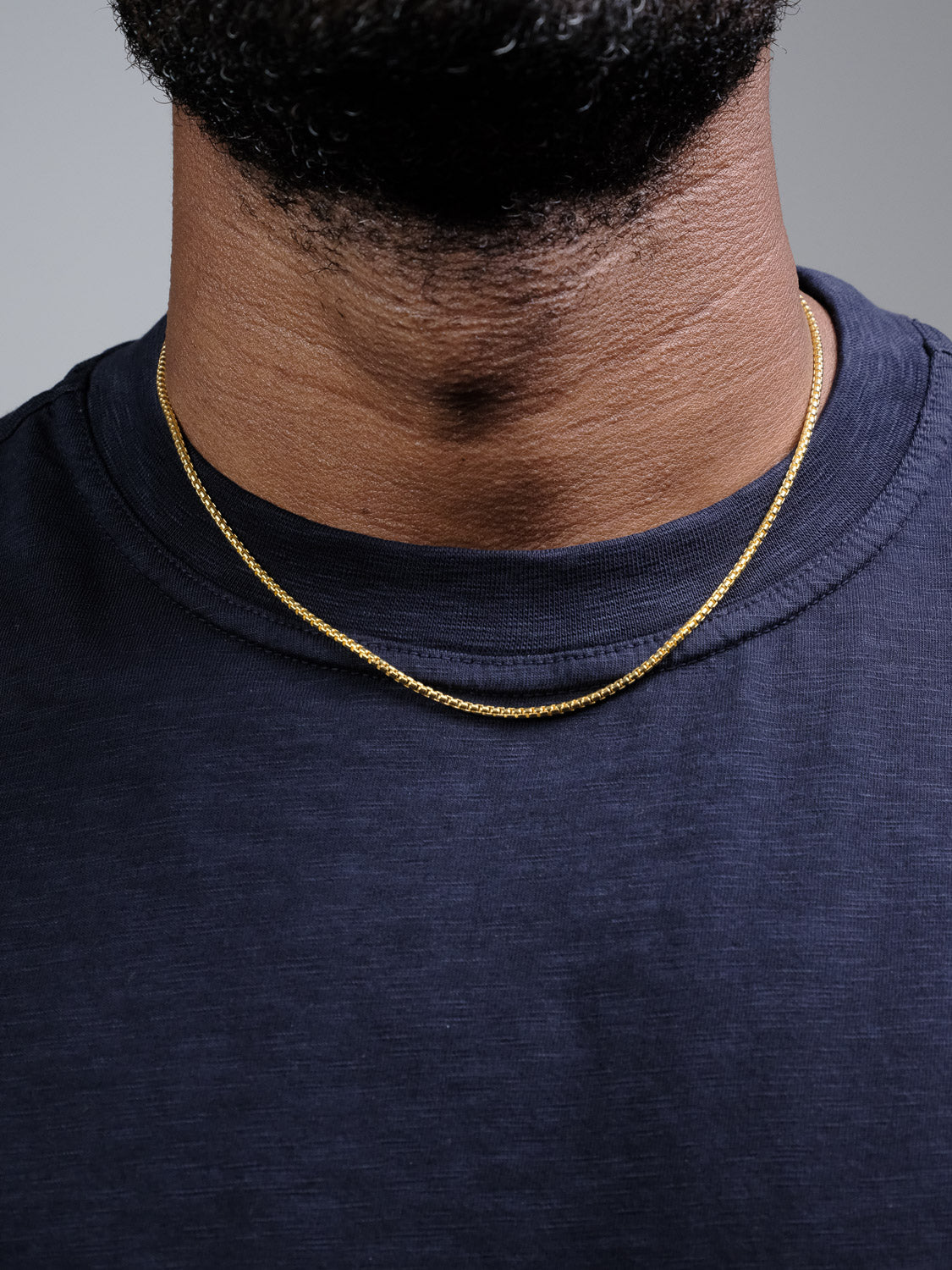 A close-up shows a person wearing the Ezra 40 Box Chain Necklace by ID Menswear and a navy blue shirt, with only the lower half of their face and neck visible.