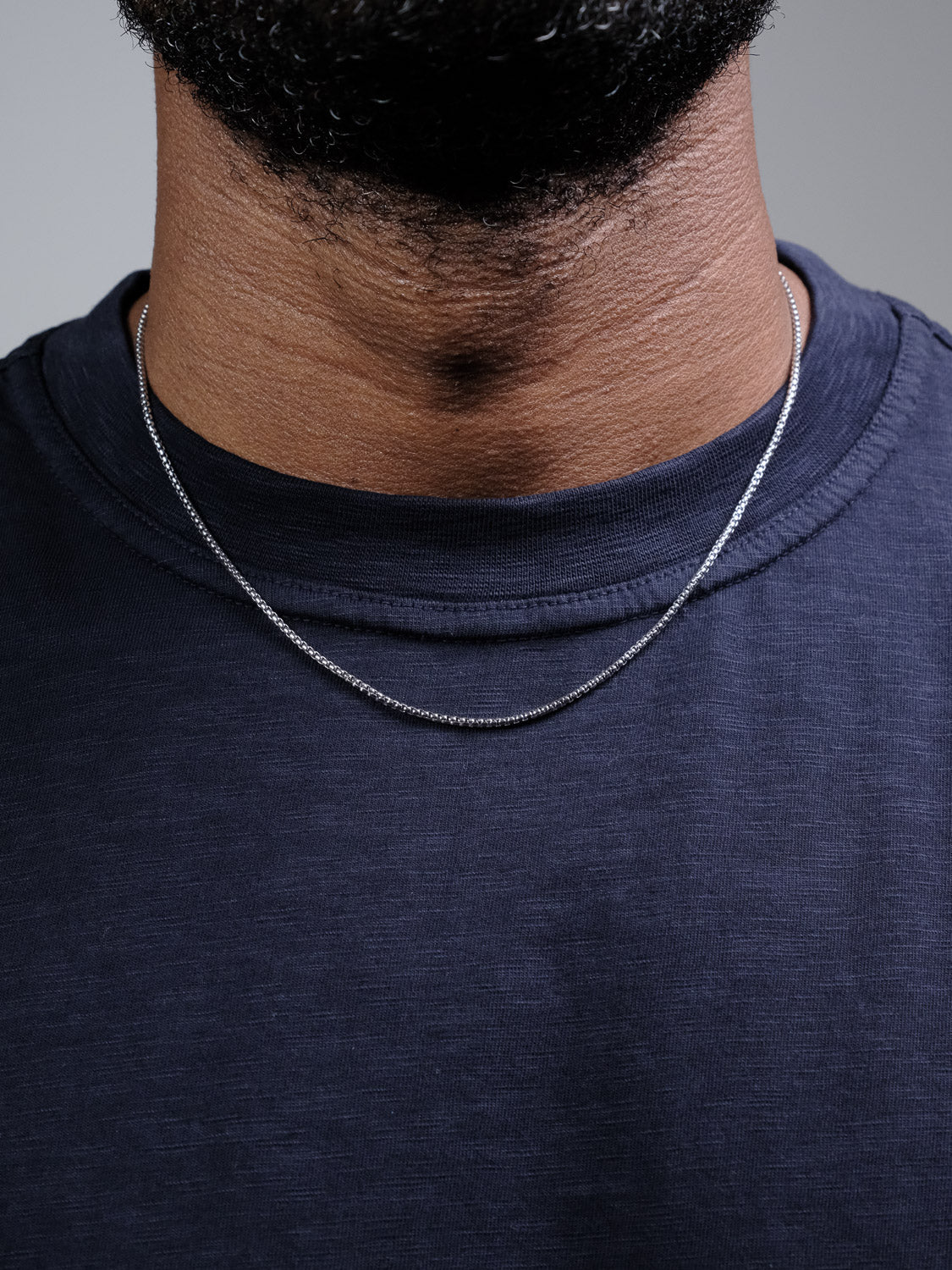 A close-up of a person’s neck wearing the Ezra 35 Box Chain Necklace by ID Menswear with a dark navy blue shirt. The 3mm sterling silver chain, made in Italy, subtly shines while the person's beard is partially visible above.