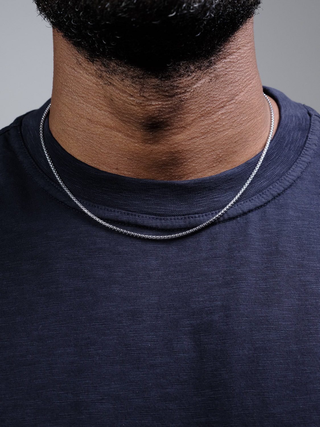 Close-up of a person wearing the Ezra 40 Box Chain Necklace by ID Menswear over a dark blue crew neck t-shirt. The thin 925 Sterling Silver chain rests on the neck, with a partial view of the lower face and beard.