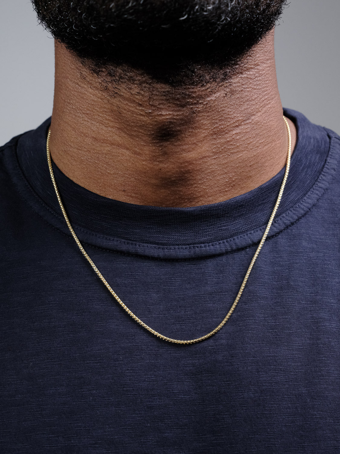 A close-up shows the Ezra 35 Box Chain Necklace by ID Menswear on a person's neck over a dark navy crew neck shirt, focusing on the jewelry and upper chest with part of the lower face slightly visible.