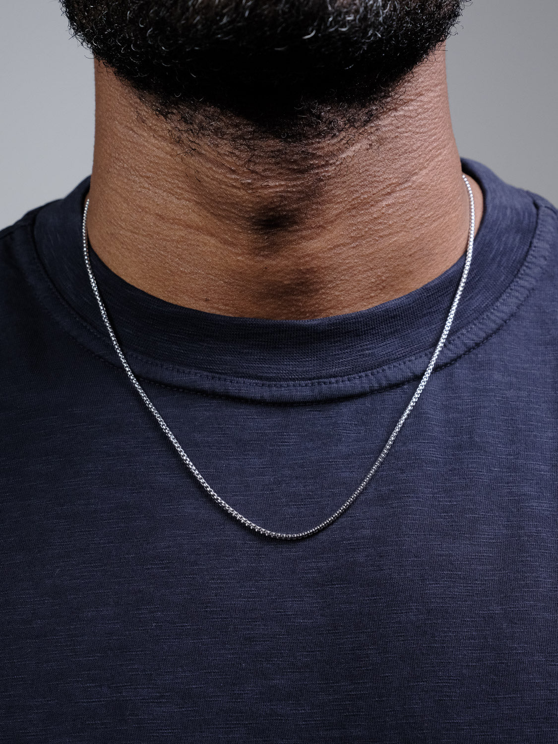 Close-up of a person wearing the Ezra 35 Box Chain Necklace by ID Menswear, a 3mm sterling silver piece made in Italy, styled with a dark navy crew-neck shirt. The focus is on the elegant chain and neck; the face is not shown.