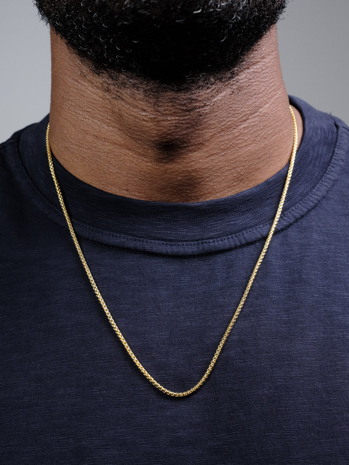 Close-up of a person wearing the Ezra 40 Box Chain Necklace by ID Menswear over a dark navy blue t-shirt, showing only the lower face, neck, and upper chest against a neutral gray background.