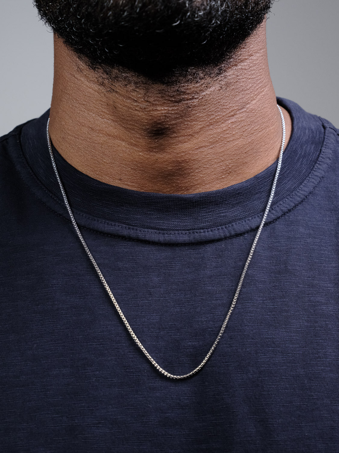 A close-up of someone wearing the Ezra 35 Box Chain Necklace by ID Menswear—a 3mm sterling silver chain made in Italy—paired with a dark blue crew-neck shirt. Only their neck and lower face are shown.
