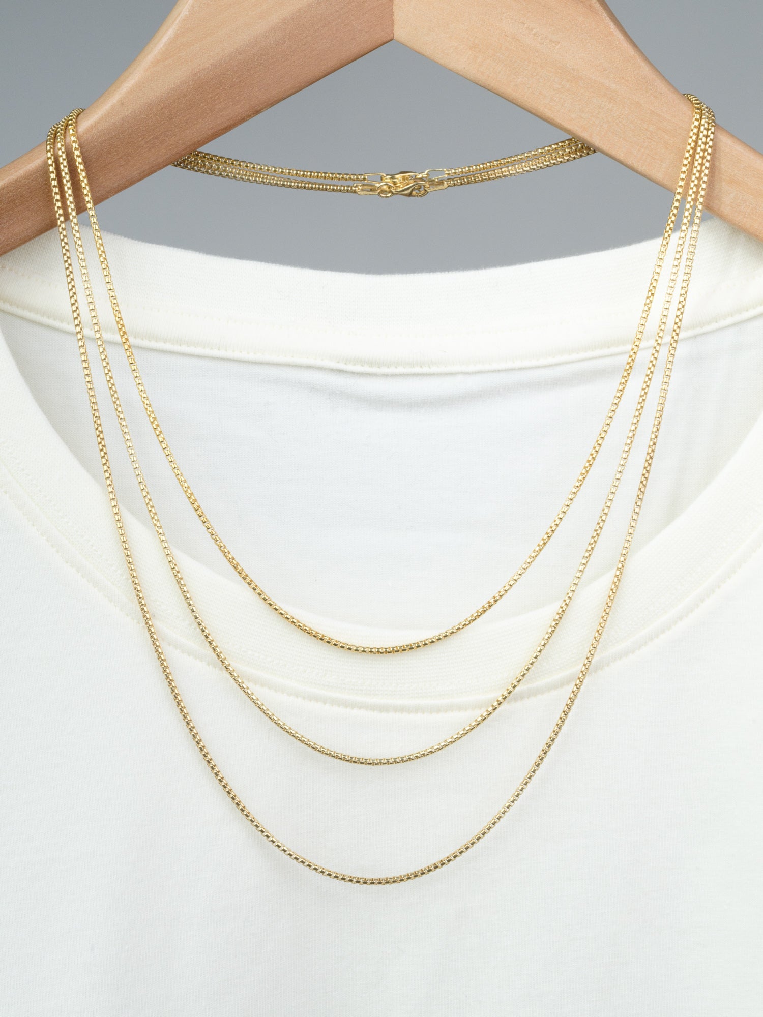 A white shirt on a wooden hanger with three thin gold plated necklaces—including the Ezra 35 Box Chain Necklace by ID Menswear—draped over it, set against a plain gray background.
