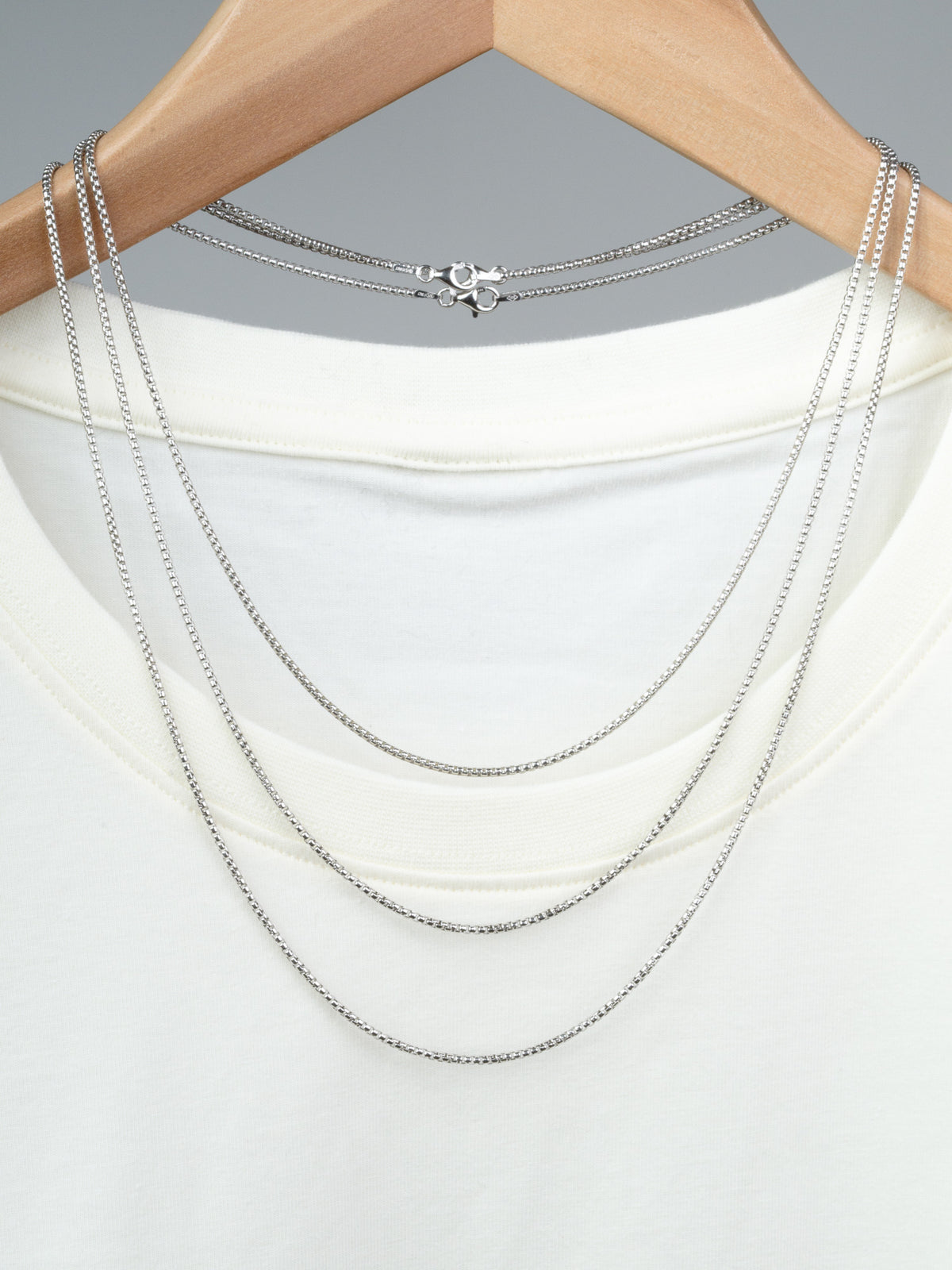 Displayed on a wooden hanger over a white t-shirt, the Ezra 35 Box Chain Necklace by ID Menswear showcases three layered sterling silver chains of varying lengths, including a delicate box chain strand.
