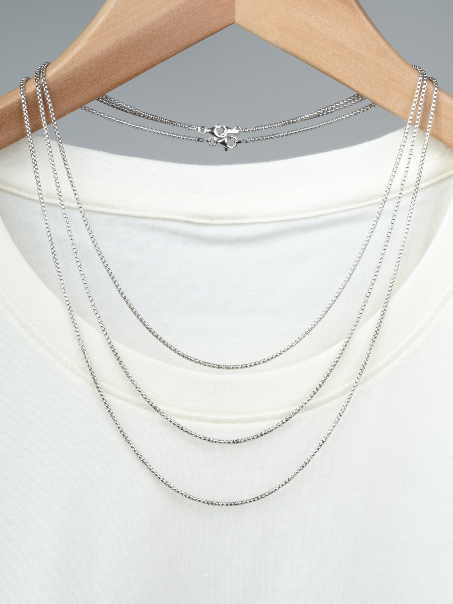 Displayed on a wooden hanger over a white t-shirt, the Ezra 35 Box Chain Necklace by ID Menswear showcases three layered sterling silver chains of varying lengths, including a delicate box chain strand.