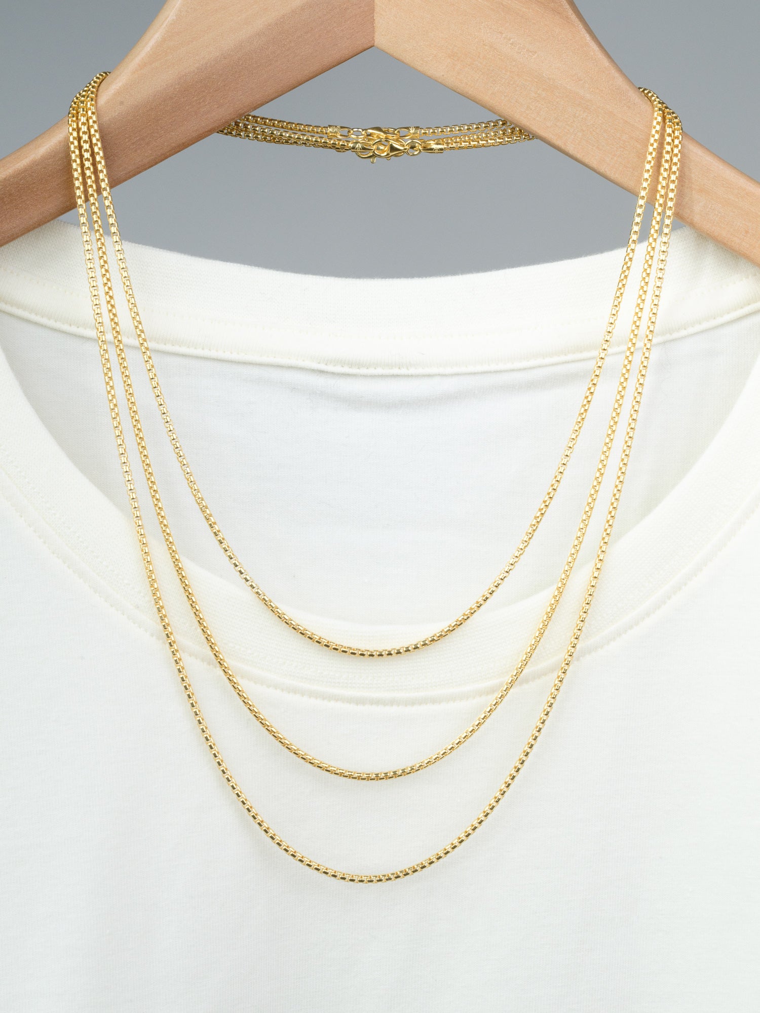 A white T-shirt on a wooden hanger with three layered gold plated necklaces, including the Ezra 40 Box Chain Necklace by ID Menswear—crafted in Italy from sleek 925 Sterling Silver—displayed against a plain gray background.