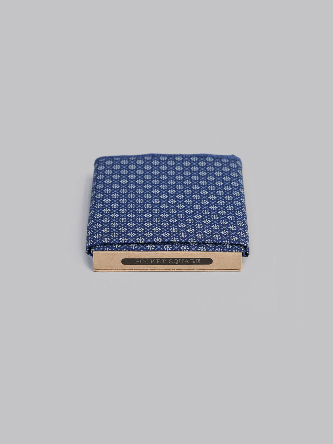 The Fleur Pocket Square by ID Menswear is a handmade blue cotton accessory with a small white geometric print, packaged in a light brown holder labeled POCKET SQUARE, shown against a plain light gray background.