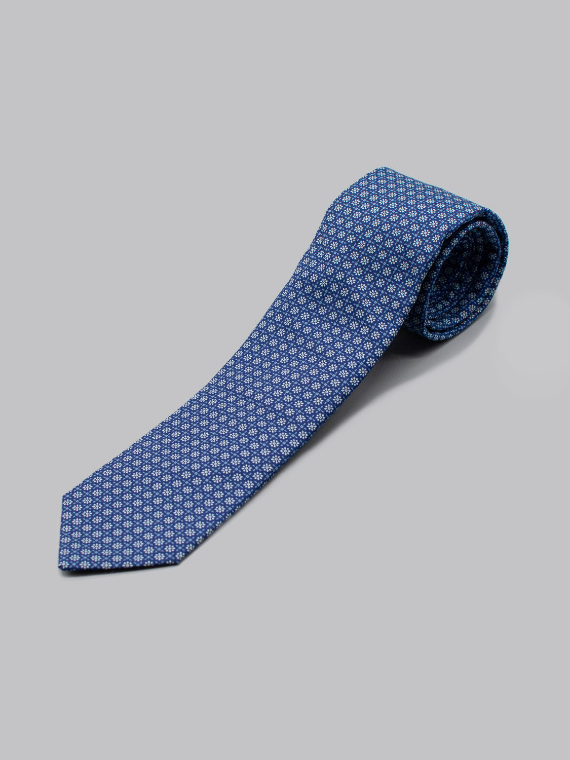 The Japanese Printed Fleur Tie by ID Menswear is handmade in Brooklyn from 100% cotton, featuring a blue geometric white pattern. Its neatly rolled on one end and set against a plain light gray background.