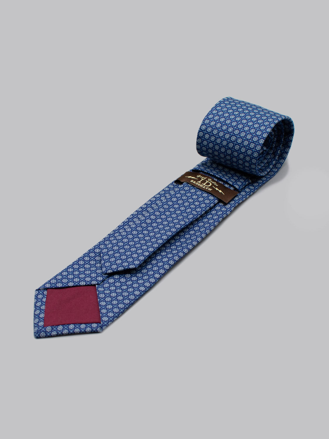 The ID Menswear Japanese Printed Fleur Tie features a blue geometric pattern, handmade in Brooklyn from 100% cotton, with a maroon inner tip accent and black label, shown on a light gray background.