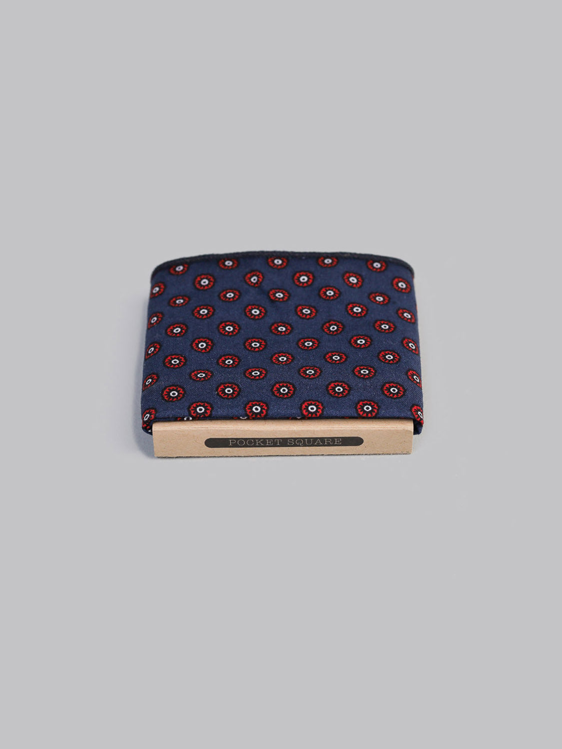 The Flodot Pocket Square by ID Menswear is a folded navy 100% cotton square, handmade in Brooklyn with a red and white circular pattern. It comes in a tan holder labeled POCKET SQUARE and is set on a plain grey background.