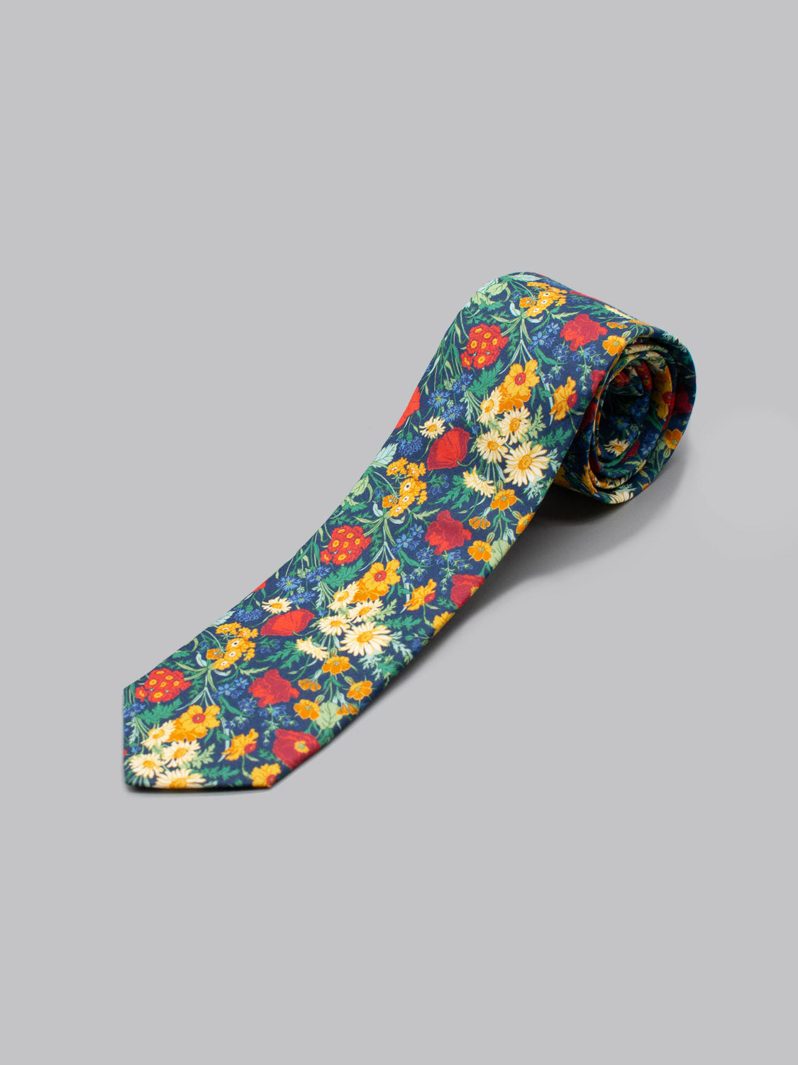 The Liberty of London Tana Lawn™ Florence tie by ID Menswear features a vibrant floral pattern with red, yellow, and white flowers on blue, handmade in Brooklyn from soft Tana Lawn cotton and shown on a plain gray surface.