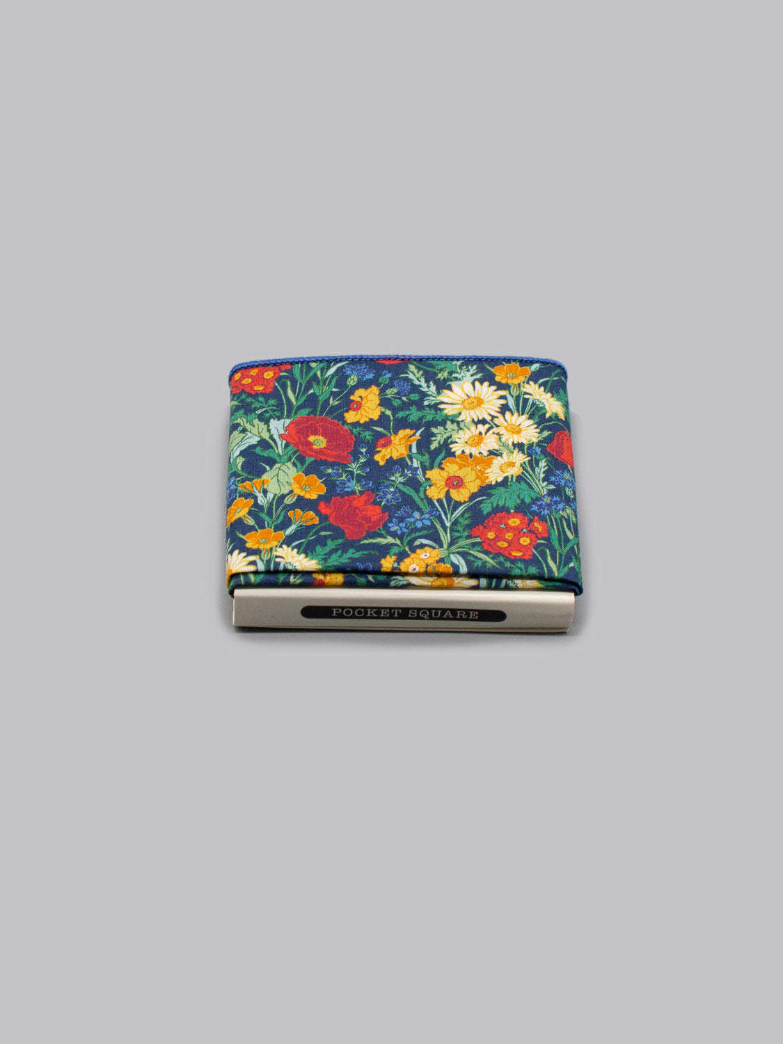 The Liberty™ Florence Pocket Square by ID Menswear, made from Tana Lawn cotton in a colorful floral print, is neatly folded and secured with a silver POCKET SQUARES clip, set against a plain gray background.